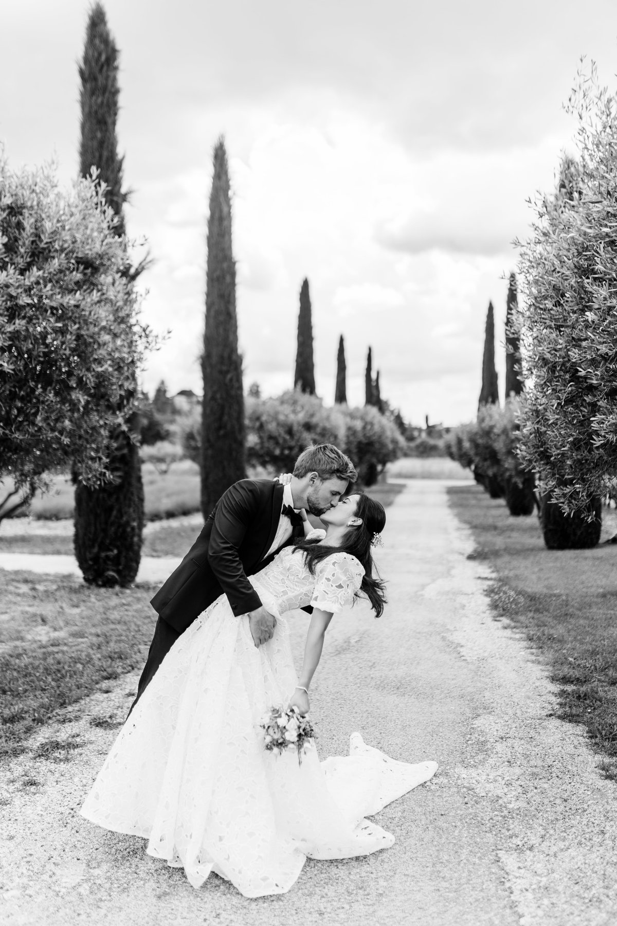 Mariés s'embrassant dans une allée de cyprès, photo de couple noir et blanc le jour du mariage